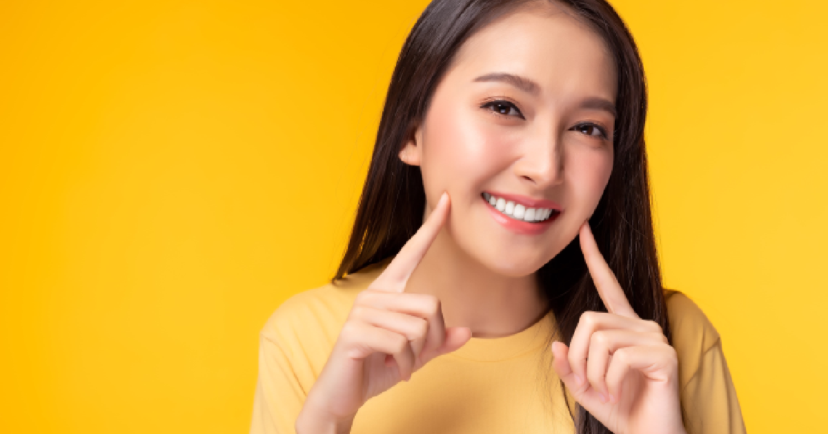 Smiling young woman with flawless white teeth pointing to her porcelain veneers, standing against a vibrant yellow background, showcasing dental aesthetics in Allentown, PA.